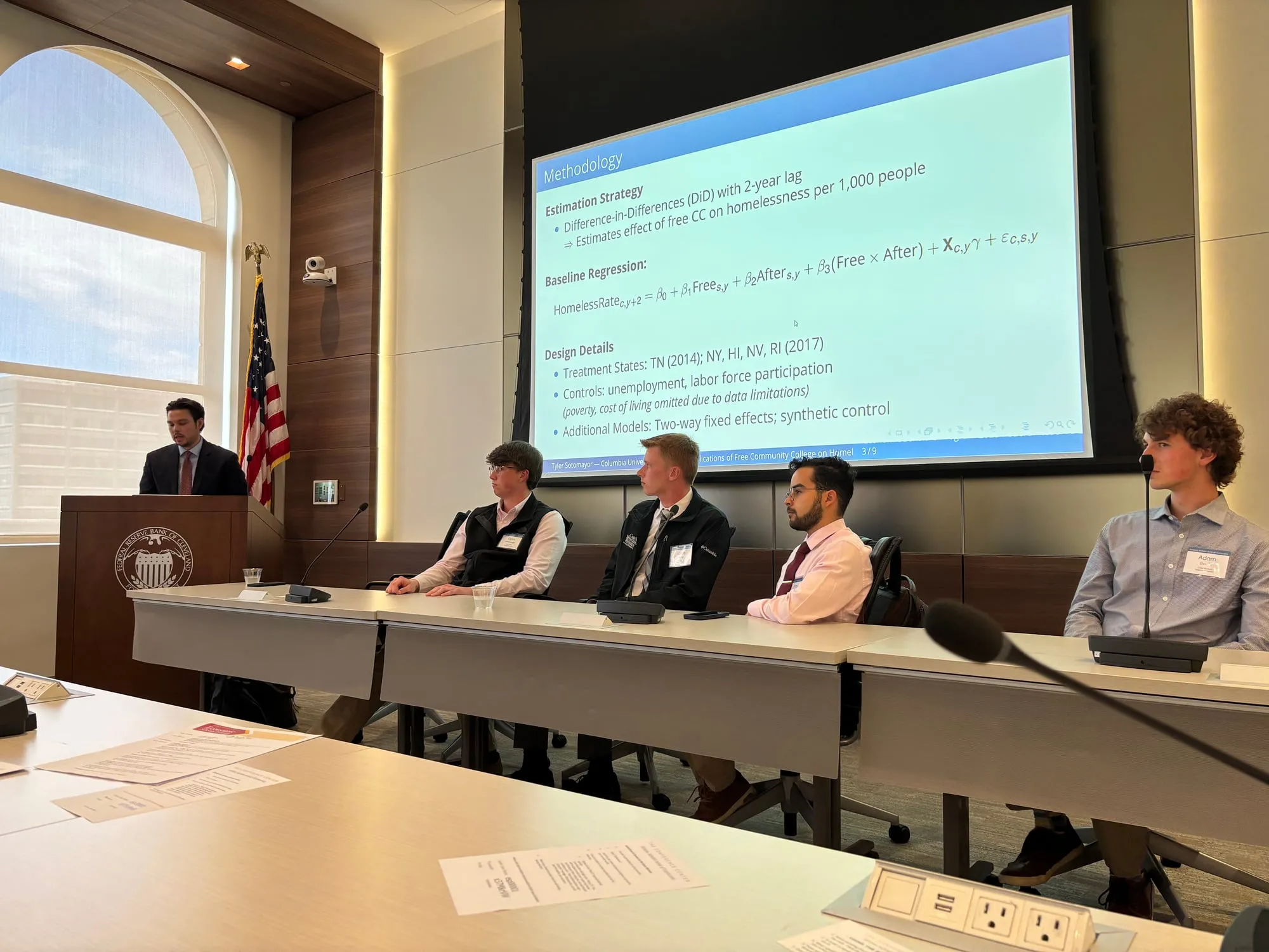 Tyler Sotomayor at the lectern discussing a panel of four student presenters at the Federal Reserve Bank of Cleveland